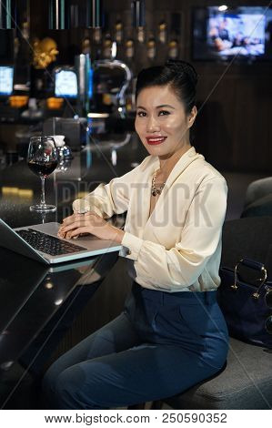Side View Of Gorgeous Happily Smiling Asian Female In Elegant Clothes Sitting At Bar Counter With Gl