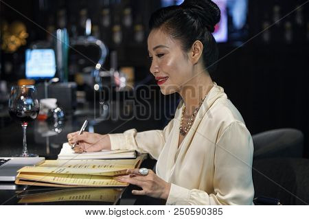 Side View Of Asian Businesswoman In Elegant Blouse Writing In Notebook Data From Bright Menu On Dark