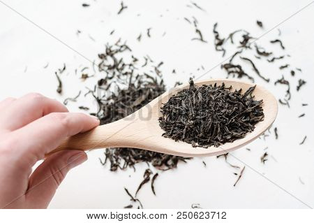Hand Holds Dry Black Tea Leaves In Wooden Spoon, Scattered Tea Leaves Over White Background, Top Vie