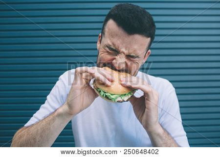 Portrait Of Man Biting A Piece Of Hamburger. He Is Shrinking. Isolated On Striped And Blue Backgroun