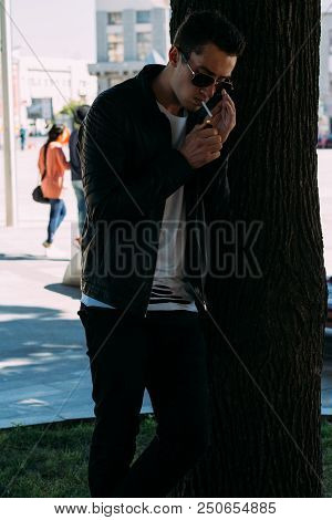 A Young Guy Smokes Near A Tree In The City. A Black Jacket, Jeans, White Sneakers, A White T-shirt A