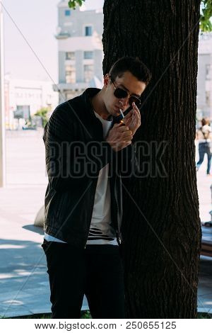 A Young Guy Smokes Near A Tree In The City. A Black Jacket, Jeans, White Sneakers, A White T-shirt A