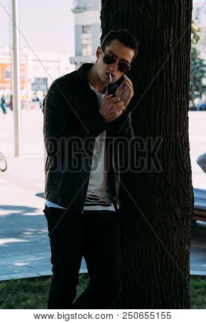A Young Guy Smokes Near A Tree In The City. A Black Jacket, Jeans, White Sneakers, A White T-shirt A