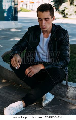 A Young Guy Smokes Near A Tree In The City. A Black Jacket, Jeans, White Sneakers, A White T-shirt A