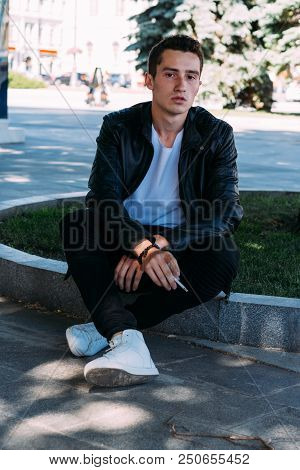A Young Guy Smokes Near A Tree In The City. A Black Jacket, Jeans, White Sneakers, A White T-shirt A