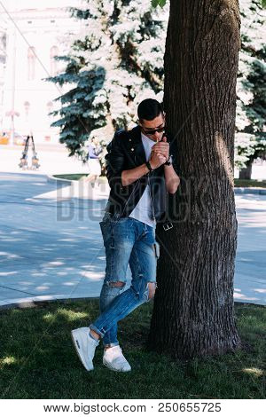 A Young Guy Smokes Near A Tree In The City. A Black Jacket, Jeans, White Sneakers, A White T-shirt A