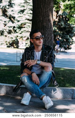A Young Guy Smokes Near A Tree In The City. A Black Jacket, Jeans, White Sneakers, A White T-shirt A
