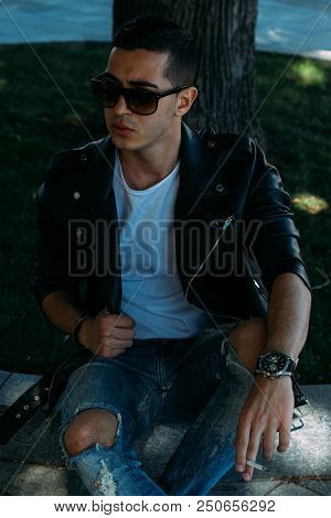 A Young Guy Smokes Near A Tree In The City. A Black Jacket, Jeans, White Sneakers, A White T-shirt A