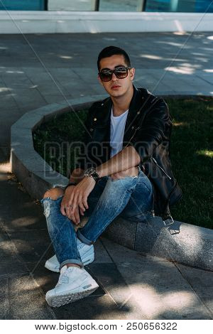 A Young Guy Smokes Near A Tree In The City. A Black Jacket, Jeans, White Sneakers, A White T-shirt A