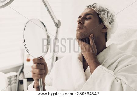 Young Handsome Man In Robe Loking Into Mirror. Smiling Man. Man In White Bathrobe. Anti-aging Cosmet