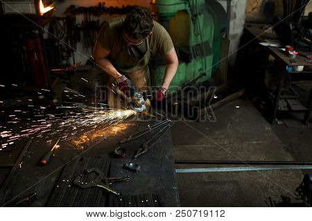 Blacksmith in workwear using electric handtool to process iron workpiece in smithy