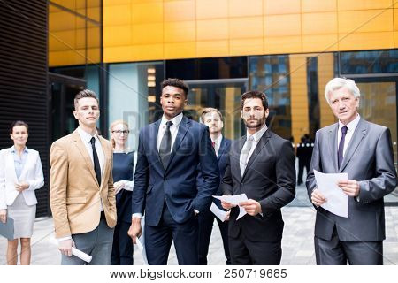 Group of confident intercultural businessmen in suits and their female colleagues standing not far from modern business center