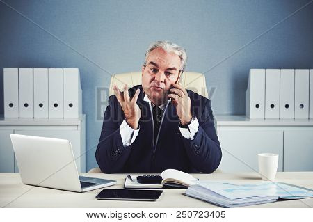 Busy Elegant Male In Dark Suit Sitting At Workplace With Gadgets And Documents And Talking On Mobile