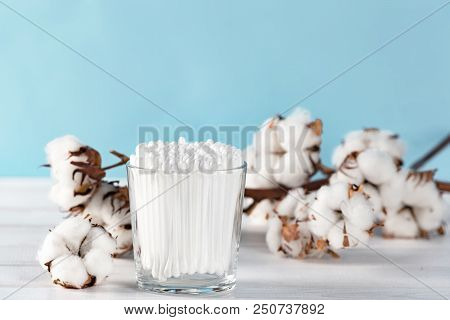 Cotton Swabs In Clear Glass Jar In Bathroom Setting With A Cotton Flowers On Pastel Blue Background.