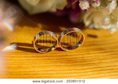 Beautiful Toned Picture With Wedding Rings Lie On A Wooden Surface Against The Background Of A Bouqu