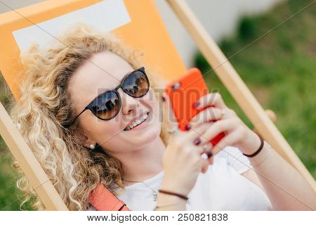 Young Curly Blonde Woman Lies In Hammock, Enjoys Summer Day, Bloggs And Chats Via Smart Phone, Wears