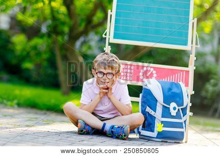 Happy Little Kid Boy With Glasses Sitting By Desk And Backpack Or Satchel. Schoolkid With Traditiona