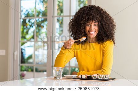 African american woman eating sushi using chopsticks at home with a happy face standing and smiling with a confident smile showing teeth