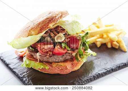 Fresh Beef Burger with Bacon, Fried Onions, Tomato Sauce, Pickled Cucumbers, Green Lettuce, Cheese and French Fries Garnish. Hamburger or Beefburger on Natural Black Stone Plate Isolated on White