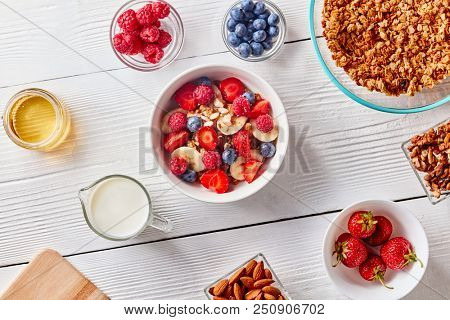 Ingredirnts for preparing healthy organic breakfast - granola, berries honey, milk, banana in white bowl on white table. Flat lay.
