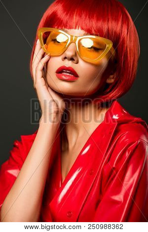 Close-up Portrait Of Young Woman In Red Latex Jacket And Vintage Yellow Eyeglasses Isolated On Grey