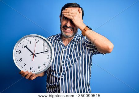 Middle age hoary man holding big clock standing over isolated blue background stressed with hand on head, shocked with shame and surprise face, angry and frustrated. Fear and upset for mistake.