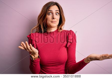 Young beautiful brunette woman wearing casual sweater stnding over pink background clueless and confused expression with arms and hands raised. Doubt concept.
