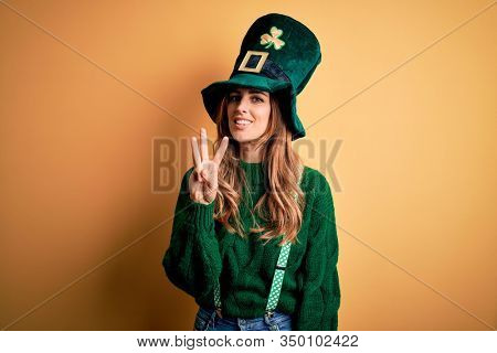 Beautiful brunette woman wearing green hat with clover celebrating saint patricks day showing and pointing up with fingers number three while smiling confident and happy.