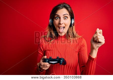 Young beautiful brunette gamer woman playing video game using joystic and headphones screaming proud and celebrating victory and success very excited, cheering emotion