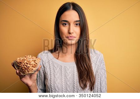 Young beautiful brunette woman holding bowl with peanuts over isolated yellow background with a confident expression on smart face thinking serious