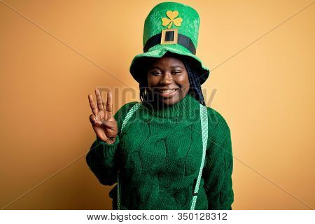 Plus size african american woman with braids wearing green hat with clover on st patricks day showing and pointing up with fingers number three while smiling confident and happy.
