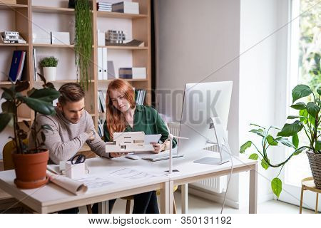Architects With Model Of House Sitting At The Desk Indoors In Office, Working.