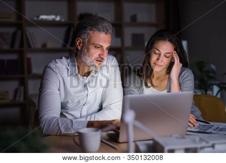 Architects With Laptop Sitting At The Desk Indoors In Office, Working Late.