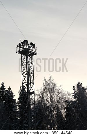 Black Silhouette Of Metal Construction With Spotlights On Top. Dark Silhouettes Of Trees On Backgrou