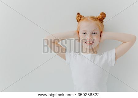 Indoor Shot Of Glad Smiling Girl Covers Ears With Hands, Ignores Noise, Has Two Ginger Buns, Freckle