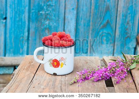Old Enamel Cup With Handful Of Fresh Rasperries And A Bunch Violet Field Flowers On Rustic Wooden Ta