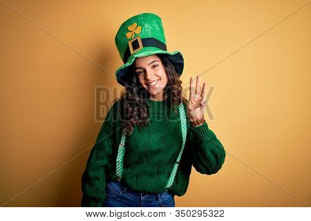 Beautiful curly hair woman wearing green hat with clover celebrating saint patricks day showing and pointing up with fingers number three while smiling confident and happy.