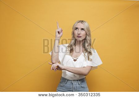 Happy Blond European Woman In White Blouse Thinking Abour Creative Idea, Pointing Up Over Yellow Bac