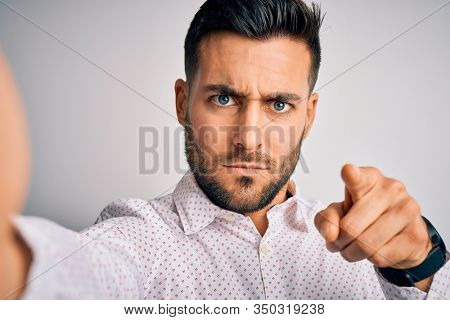 Young handsome man wearing shirt making selfie by the camera over white background pointing with finger to the camera and to you, hand sign, positive and confident gesture from the front