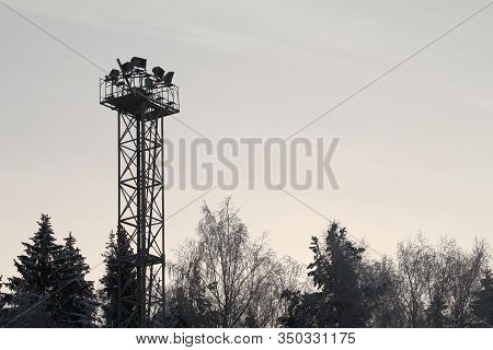 Black Silhouette Of Metal Construction With Spotlights On Top. Dark Silhouettes Of Trees On Backgrou