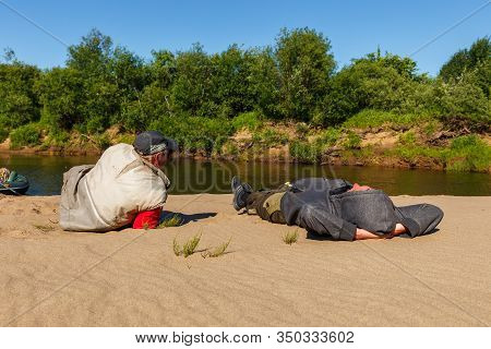 Tuzha, Russia - June 13, 2019: Two Fishermen Are Lying On The Sandy Shore, Rest On The River. Pizhma
