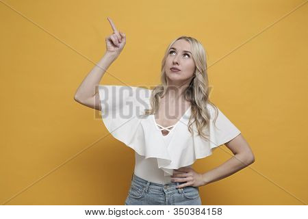 Happy Blond European Woman In White Blouse Thinking Abour Creative Idea, Pointing Up Over Yellow Bac