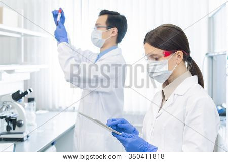 Two professional pharmacologists wearing protective masks, gloves and eyeglasses working on medicaments in modern laboratory