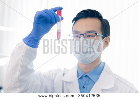 Unrecognizable Asian pharmacologist wearing eyeglasses, protective mask and gloves holding test tube with red liquid
