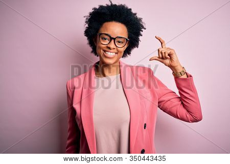 Young beautiful African American afro businesswoman with curly hair wearing pink jacket smiling and confident gesturing with hand doing small size sign with fingers looking and the camera. Measure