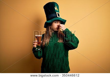 Young beautiful woman wearing green hat drinking glass of beer on saint patricks day smelling something stinky and disgusting, intolerable smell, holding breath with fingers on nose. Bad smell