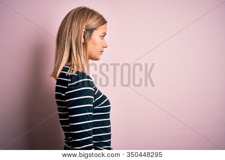 Young beautiful blonde woman wearing casual striped sweater over pink isolated background looking to side, relax profile pose with natural face with confident smile.