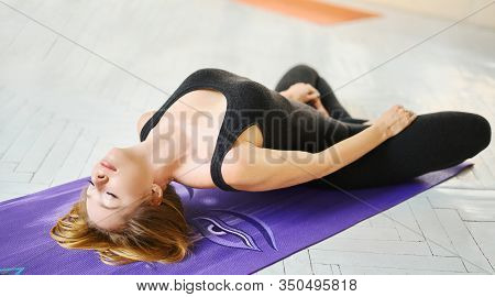 Sporty Blonde Woman In Black Sportswear Practicing Yoga, Making Stretching On Mat In Yoga Studio