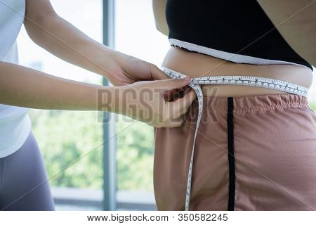 Young Female Trainer Measuring Fat Layer Of Overweight Woman With Waistline At Fitness