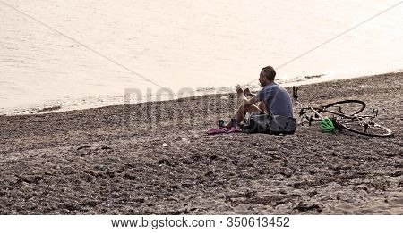 Rodby, Denmark - July 19, 2019: A Man Sitting On The Beach With His Mobile Phone At Sunset
